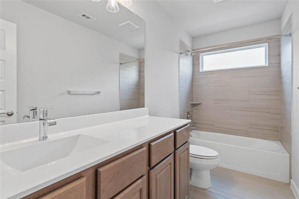 Secondary bath features tile flooring, bath/shower combo with tile surround, stained wood cabinets, beautiful light countertops, mirror, dark, sleek fixtures and modern finishes.