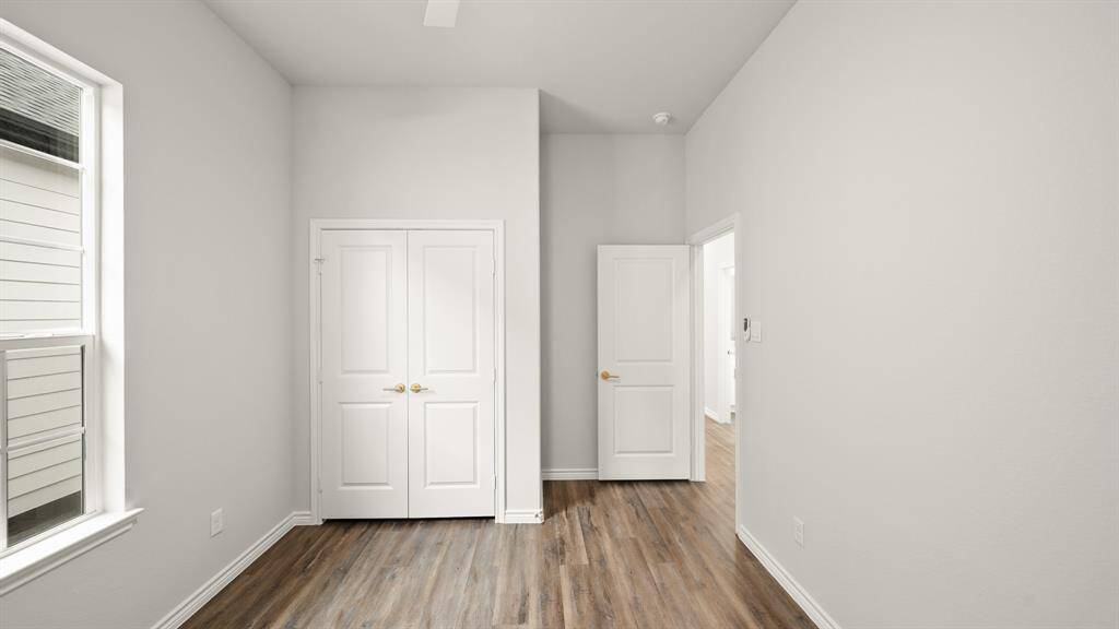 Bright bedroom with light gray walls, wood flooring, and a large window. It features a double-door closet and an additional door leading to another area.