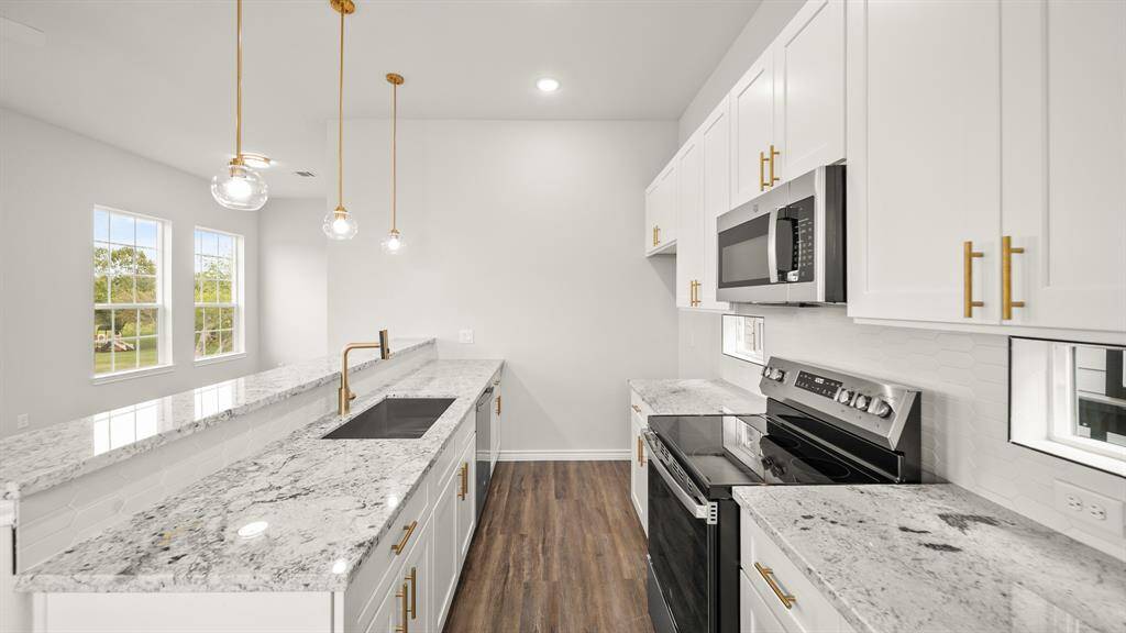 This kitchen features modern white cabinetry with gold handles, sleek granite countertops, and stainless steel appliances. The space is brightened by pendant lights and natural light from large windows.