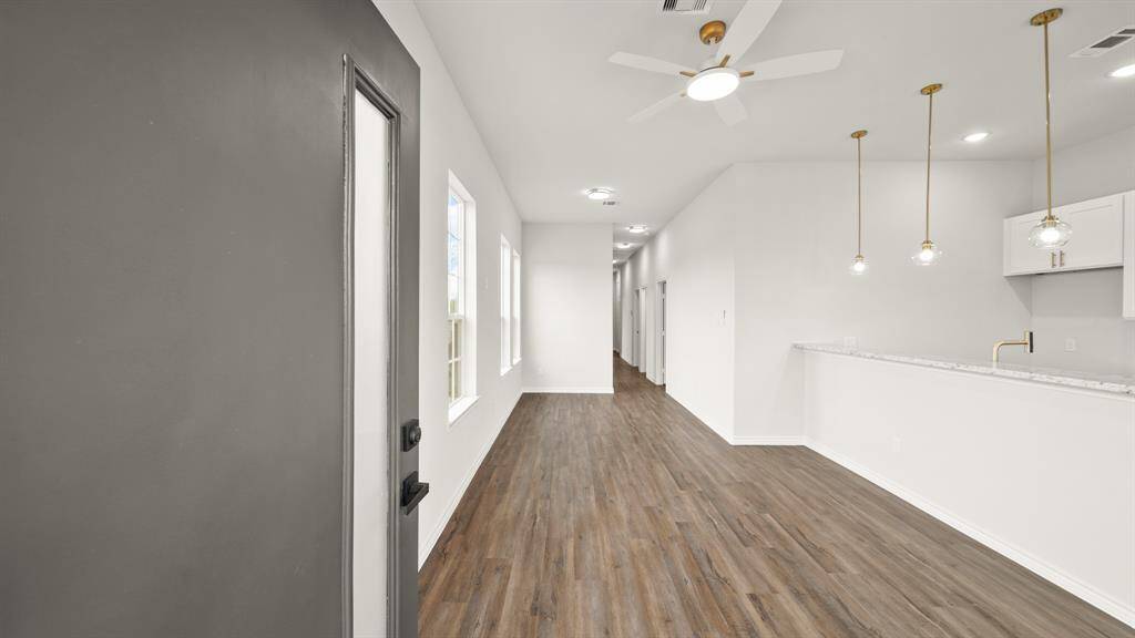 Modern open-concept entryway and living area with wood flooring and neutral walls. It features a ceiling fan, pendant lighting, and a kitchen bar with white cabinets, offering a clean and spacious look.