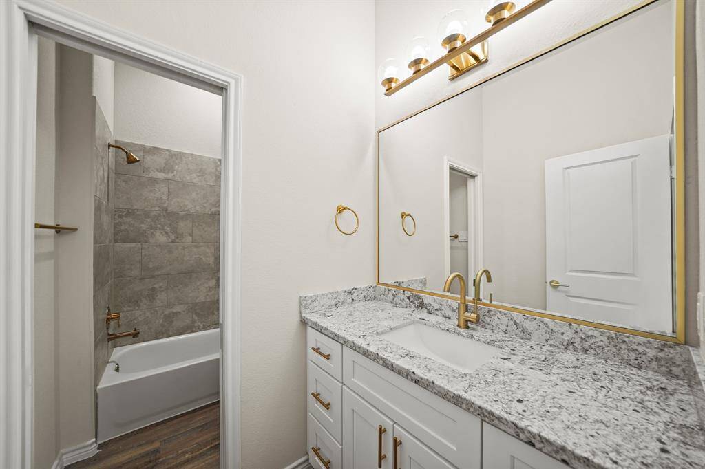 This modern bathroom features a sleek granite countertop with a large mirror and gold fixtures. It includes a clean white vanity with ample storage and a bathtub with neutral-toned tile surround, providing a stylish and functional space.