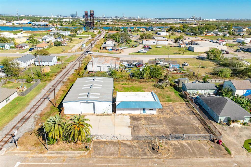 This aerial photo features a property with a large industrial building and paved area, situated near a railway track. The neighborhood includes a mix of residential and commercial structures, with visible greenery and distant industrial facilities.