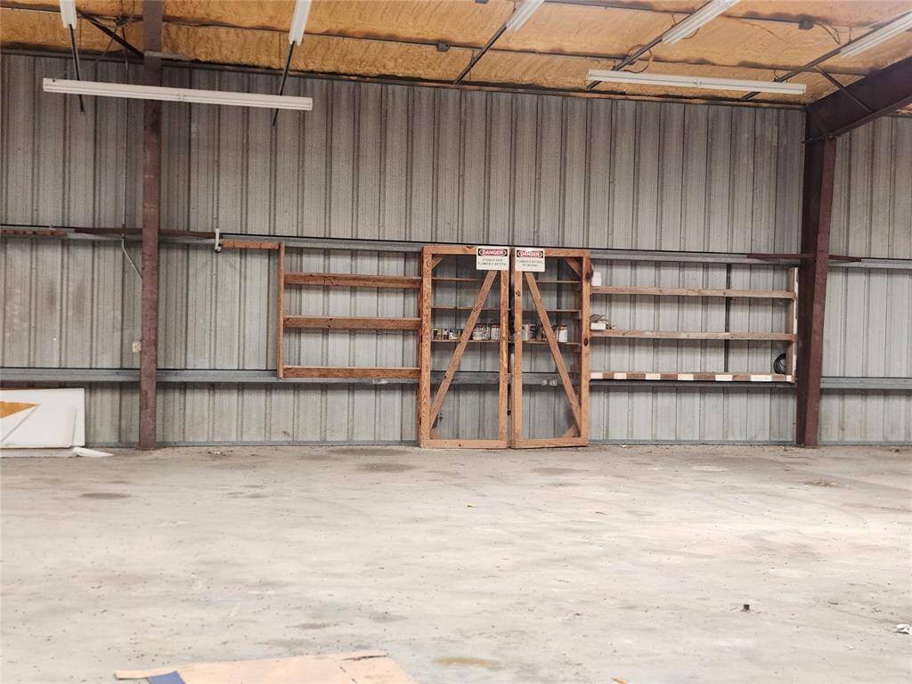 This photo shows a spacious, industrial-style garage or workshop area with a high ceiling. It features metal siding, exposed beams, and built-in wooden shelving, suggesting ample storage and workspace potential. Ideal for projects or storage.