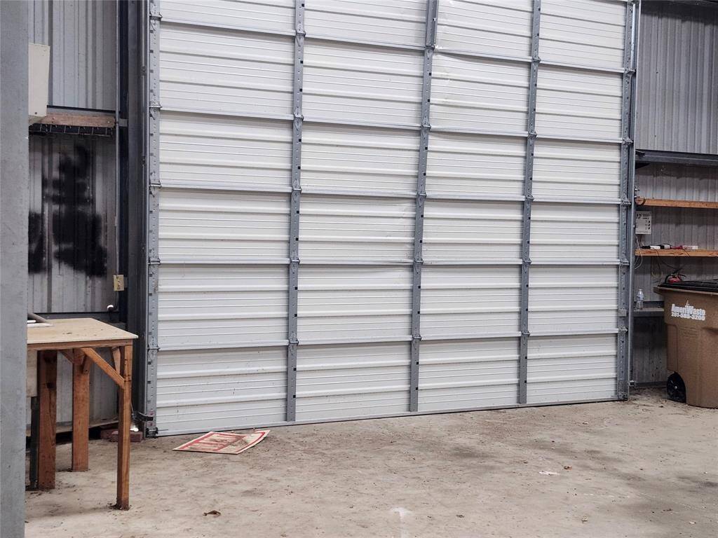 This photo shows the interior of a spacious garage or workshop, featuring a large metal roll-up door. The space has a concrete floor and some shelving, suitable for storage or projects.