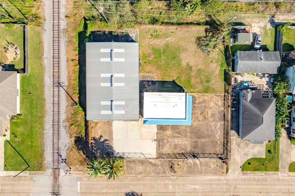 This aerial photo showcases a property with a large metal-roofed building and ample paved space, situated next to a railroad track. The lot includes grassy areas and is bordered by residential homes, offering potential for various uses.