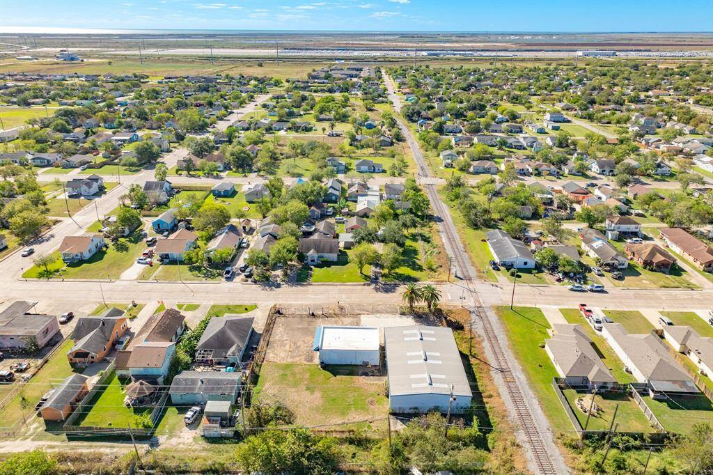 An aerial view of a residential neighborhood with neatly arranged houses, green lawns, and tree-lined streets. A railway track runs through the area, and there's a mix of open spaces and developed land. The horizon shows a vast expanse, giving a sense of openness.