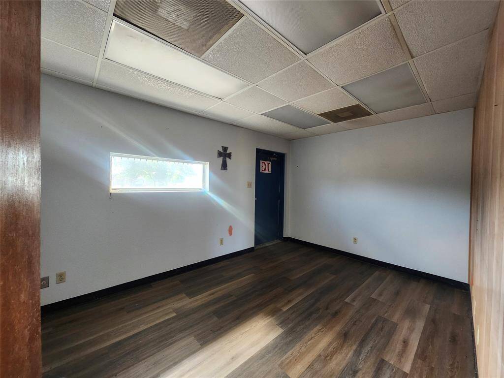 A small room with laminate wood flooring and a drop ceiling. It has a narrow window, an exit door, and plain walls, offering potential for customization.