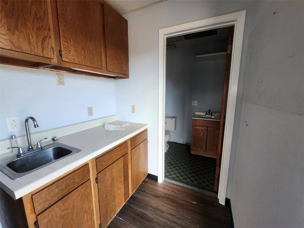 This photo shows a small kitchen area with wooden cabinets, a sink, and a counter. Adjacent to it is a bathroom with a toilet and sink, featuring outdated flooring. The space appears to need some updating or renovation.