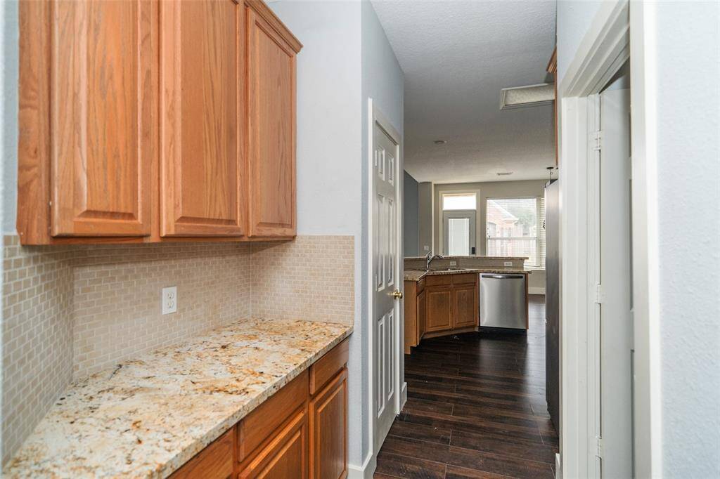 This photo shows a hallway leading to a kitchen area. On the left, there are wooden cabinets and a granite countertop. The kitchen features more wooden cabinetry, stainless steel appliances, and dark hardwood flooring. Natural light comes through a window and glass door at the far end.