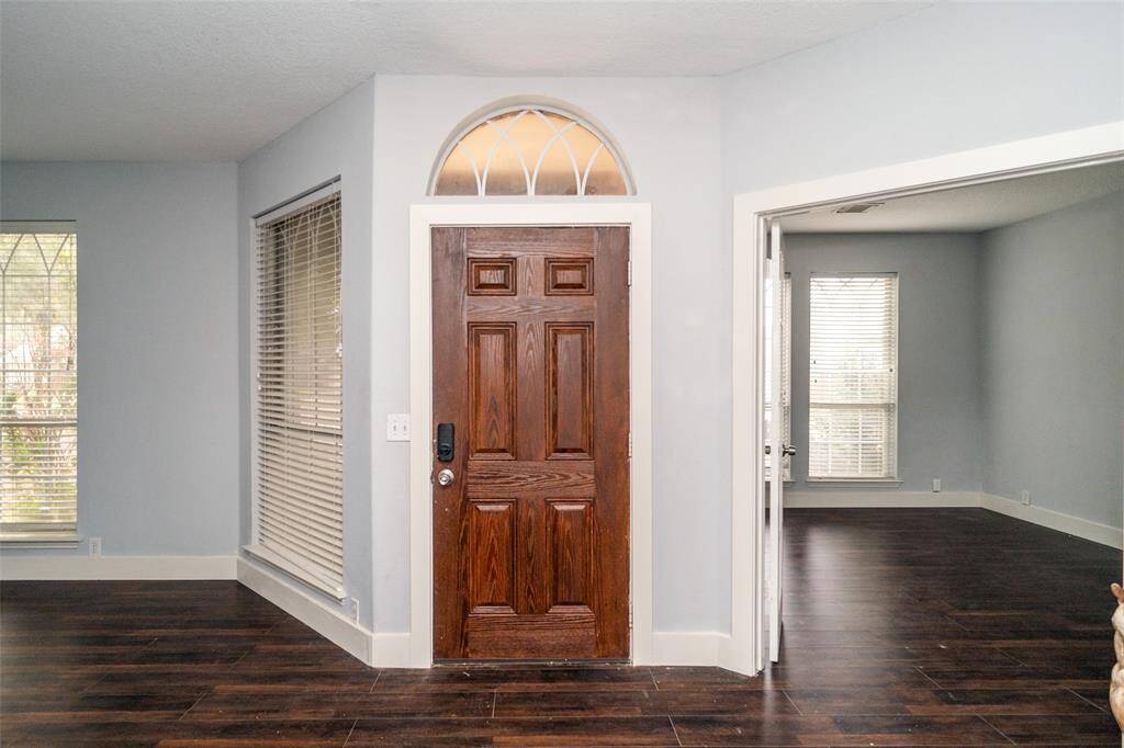 Bright entryway with a wooden door and transom window, connecting to a spacious room with large windows, offering plenty of natural light and dark wood flooring.