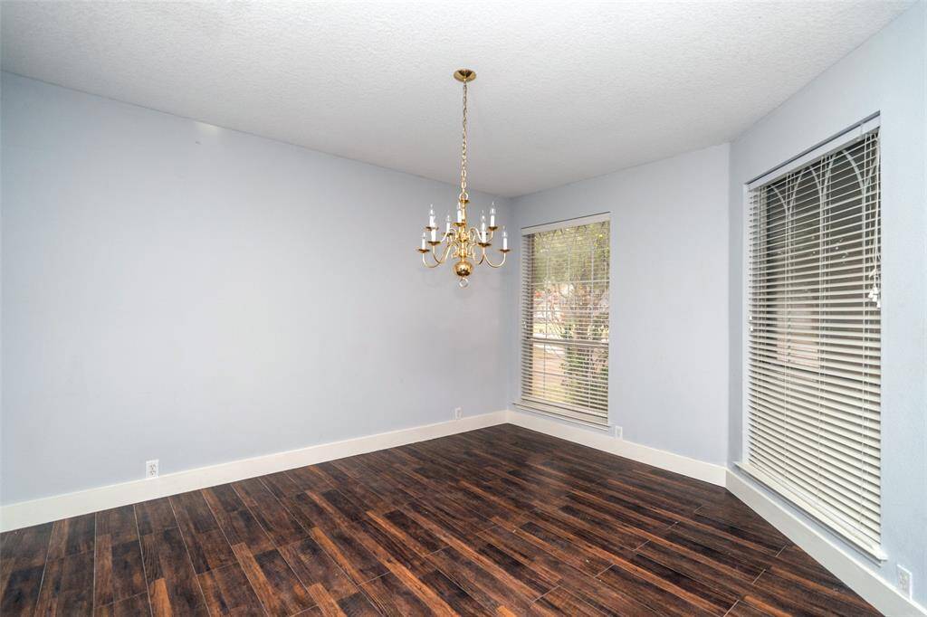 This room features elegant dark wood flooring, soft gray walls, and a classic gold chandelier. Large windows with blinds provide ample natural light. Ideal for a dining or living space.