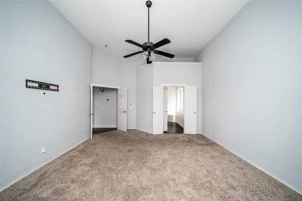 This spacious room features high ceilings, neutral carpeting, and a ceiling fan. It includes a double-door entry to an adjoining space with ample natural light.