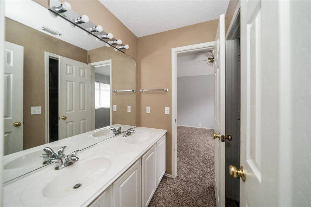 This photo shows a bathroom with a double vanity and large mirror, leading into a carpeted bedroom. The space has neutral tones and modern fixtures, offering a clean and functional layout.