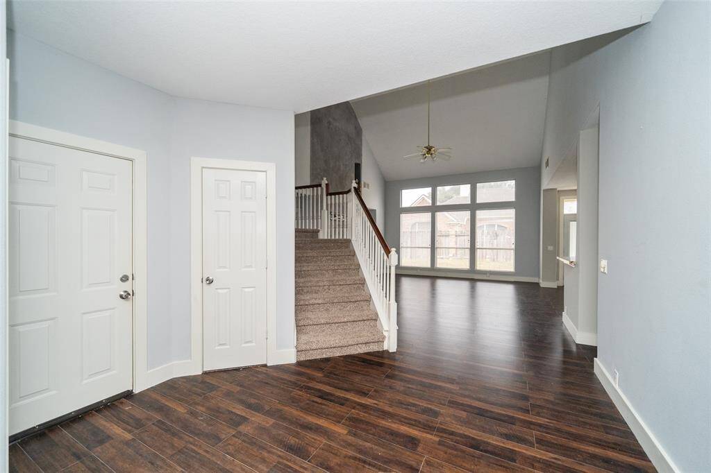 This welcoming entryway features elegant dark wood flooring and soft gray walls. It leads to a spacious living area with high ceilings and large windows that provide ample natural light. A staircase with carpeted steps adds a cozy touch.