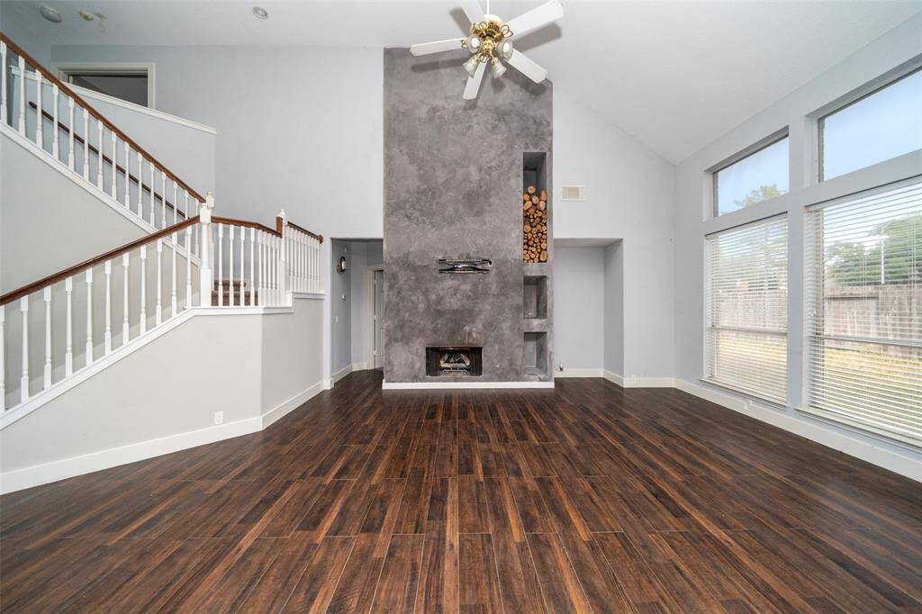 This bright living room features a high ceiling, large windows, and a modern fireplace with a wood storage niche. It boasts dark hardwood floors and an elegant staircase, creating a spacious and stylish atmosphere.