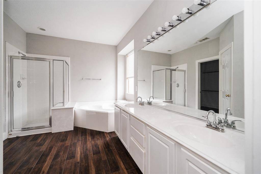This bathroom features a spacious layout with a large mirror, dual sinks, a glass-enclosed shower, and a corner bathtub. The room is enhanced by wood-style flooring and ample natural light.