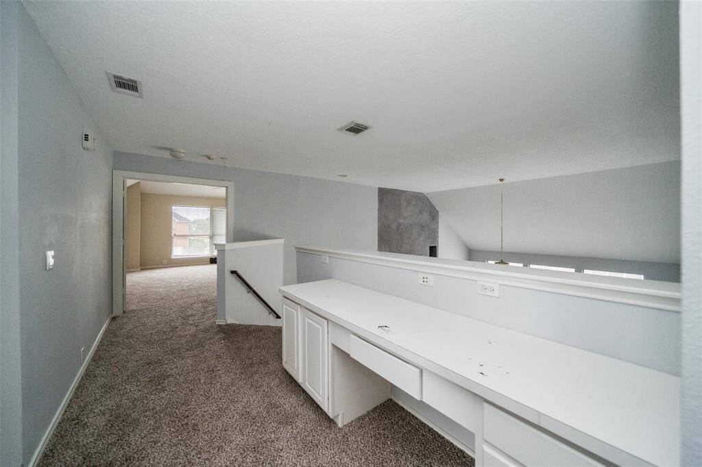This photo shows a spacious upstairs landing with a built-in desk and storage, carpeted flooring, and a view into a well-lit room with large windows.