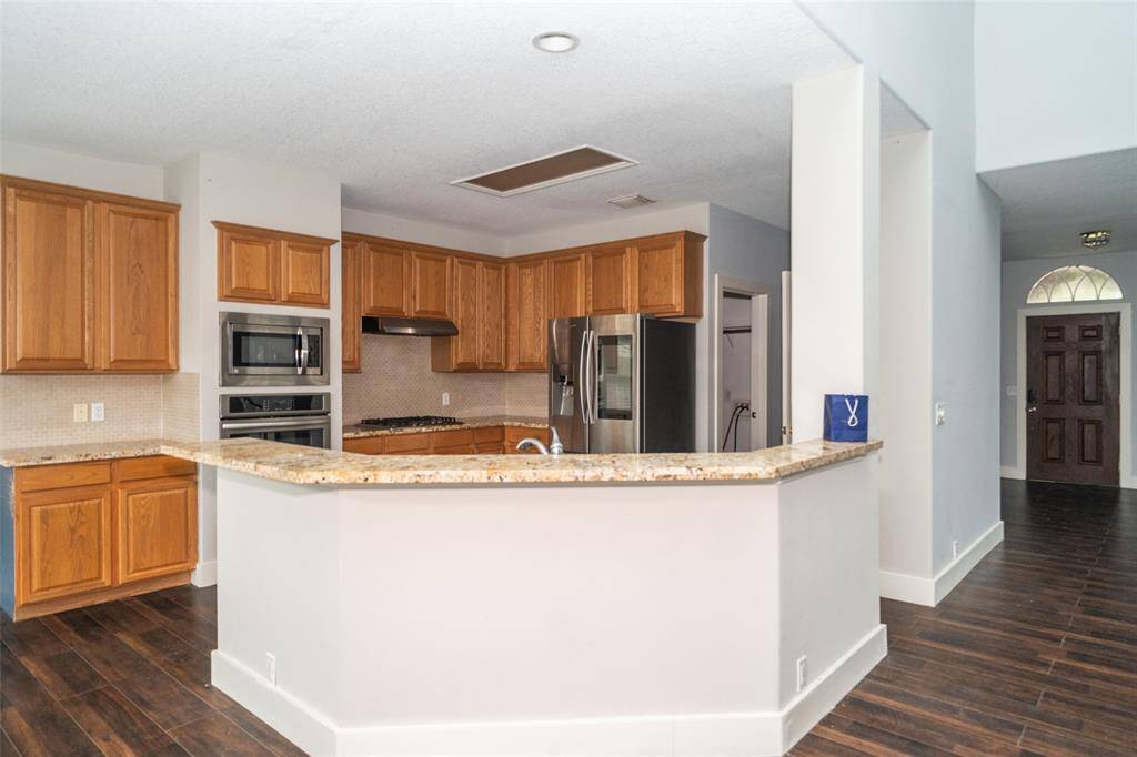 This kitchen features a spacious layout with granite countertops and warm wooden cabinetry. It includes stainless steel appliances, a built-in microwave and oven, and a large fridge. The open design seamlessly connects to the living area, complemented by dark hardwood flooring.