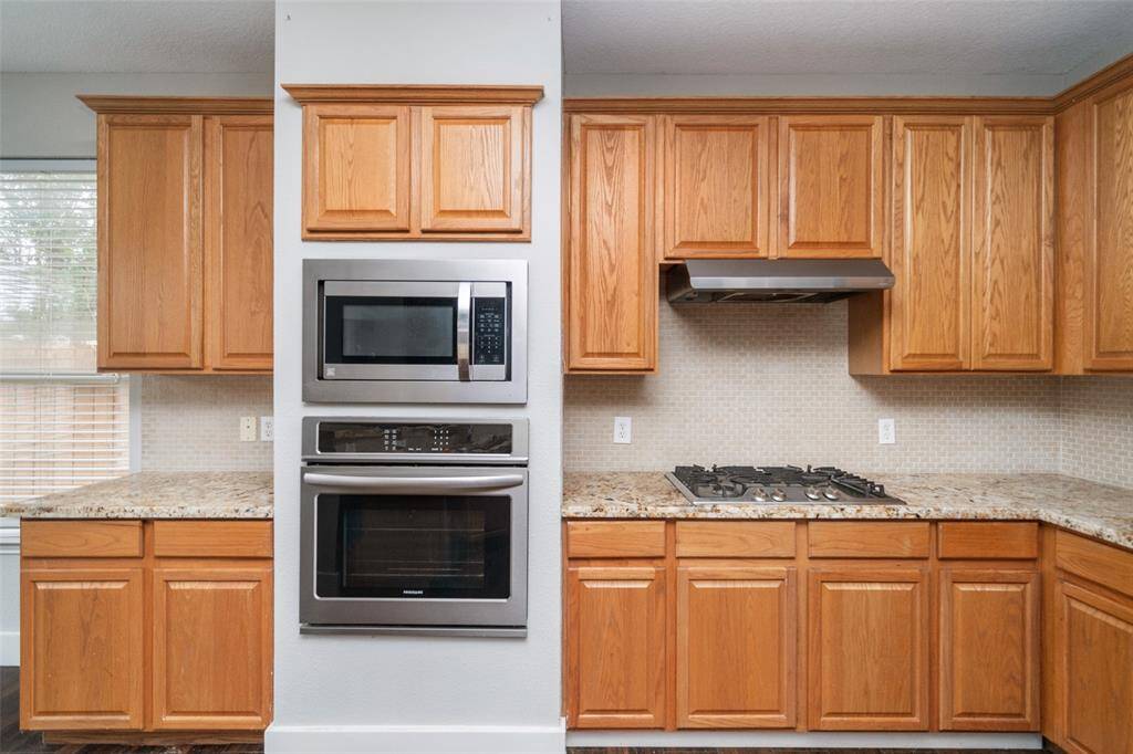 This kitchen features wooden cabinetry, granite countertops, a modern gas cooktop with a stainless steel hood, and built-in microwave and oven. The neutral backsplash and ample storage make it both functional and stylish.