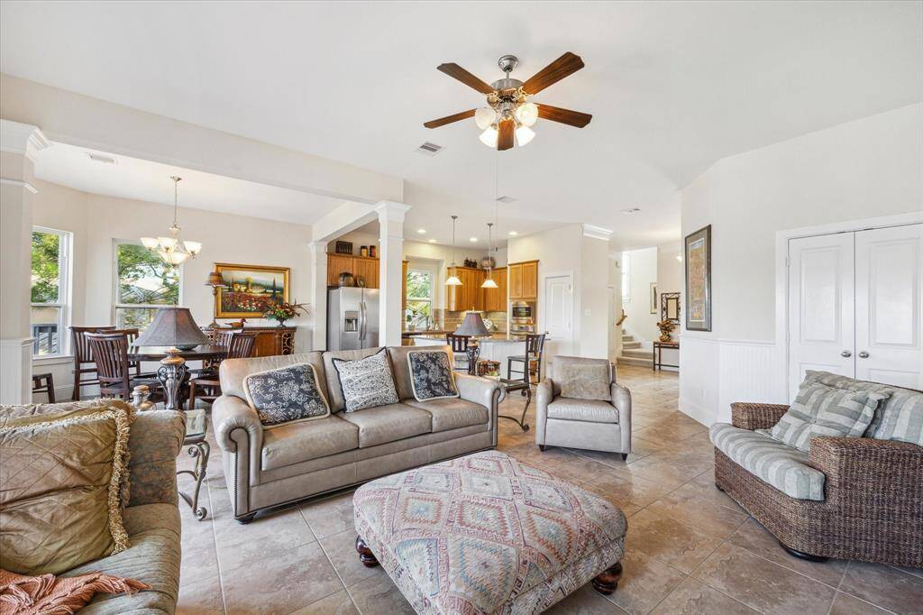 This spacious and inviting living area features high ceilings, a ceiling fan, and abundant natural light streaming through large windows. The neutral-toned tile floors, crown molding, and elegant furnishings create a comfortable and stylish atmosphere. The adjoining dining area, framed by architectural columns, offers a perfect space for gatherings or intimate dinners.