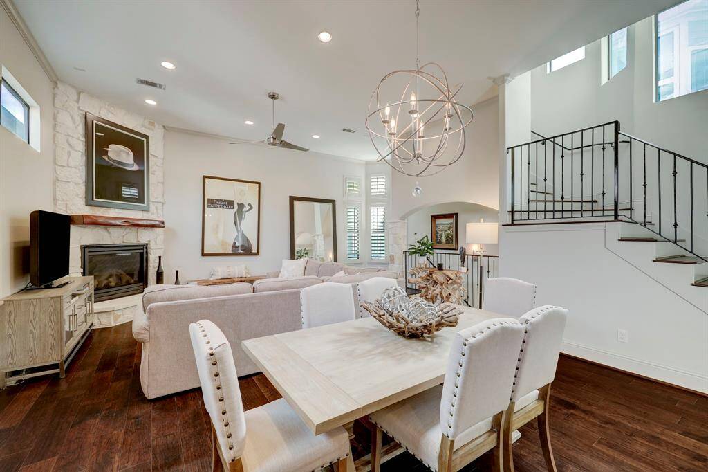 This inviting living space features an open floor plan with a cozy alamo stone fireplace, 12FT high ceilings, and modern light fixtures. The dining area blends seamlessly with the living room, highlighted by a stylish staircase and large windows that allow natural light to fill the room.