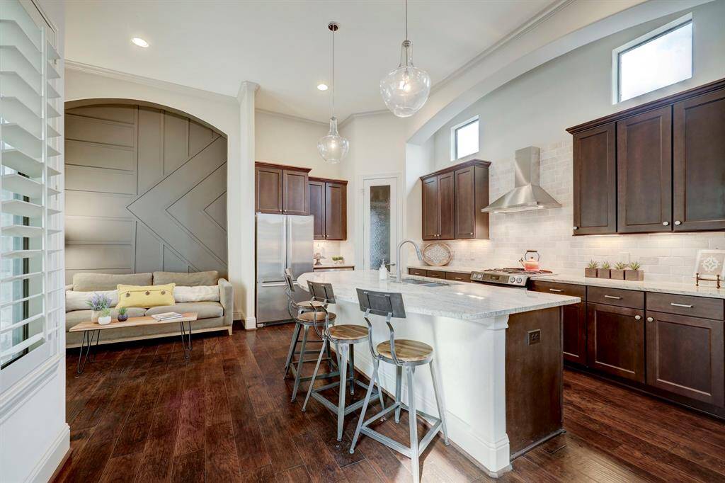 This modern kitchen features dark wood cabinets, stainless steel appliances, and a spacious island with bar seating. It opens to a cozy nook with a stylish accent wall and a comfortable sofa, all set on warm hardwood floors.