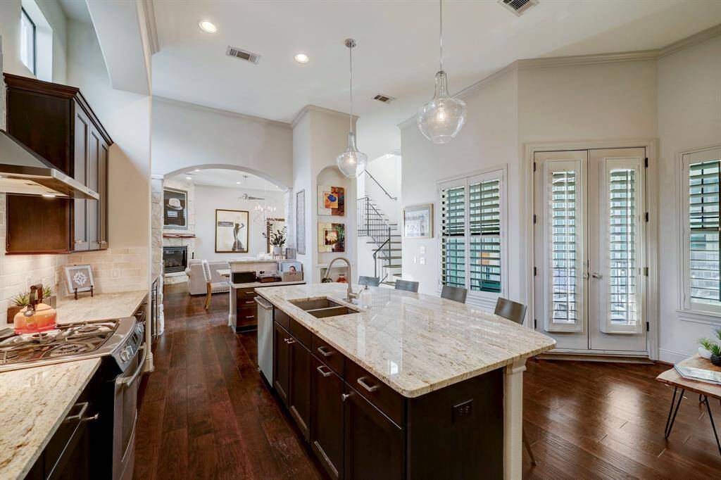 This photo features a modern, open-concept kitchen and living area. It includes a large kitchen island with granite countertops, dark wood cabinets, and stainless steel appliances. The space is well-lit with pendant lights and natural light from French doors with shutters. The living area has a cozy fireplace and hardwood floors throughout.