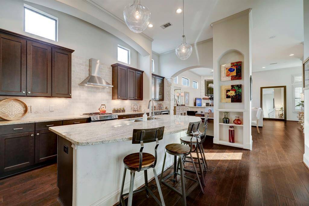 This modern kitchen features dark wood cabinets, a large marble island with seating, and stainless steel appliances. The space is bright with high ceilings, recessed lighting, and an open layout connected to the dining area. Elegant built-in shelves display decorative items.