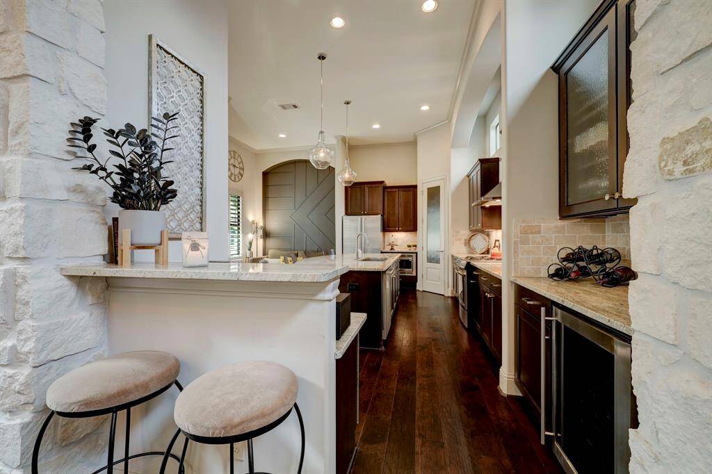 This kitchen features a modern design with dark wood cabinets, light stone countertops, and stainless steel appliances. It includes a central island with pendant lighting and a breakfast bar with seating. The space is accentuated by stone walls and hardwood floors, creating a warm and inviting atmosphere.