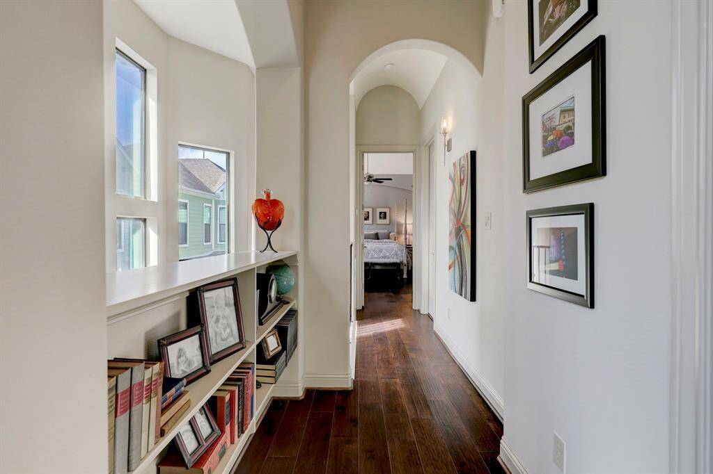This photo showcases a bright hallway with hardwood floors, featuring built-in shelves filled with books and framed photos. It leads to a cozy bedroom with a ceiling fan, creating a warm and inviting atmosphere.