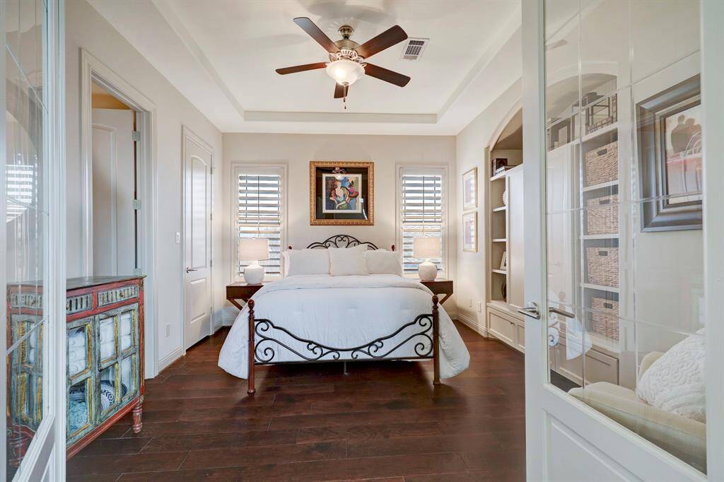 This cozy bedroom features elegant dark wood flooring, a stylish ceiling fan, and ample natural light from two windows with shutters. The room is tastefully decorated with a framed artwork above the bed, built-in shelving, and a decorative cabinet, creating a warm and inviting atmosphere.