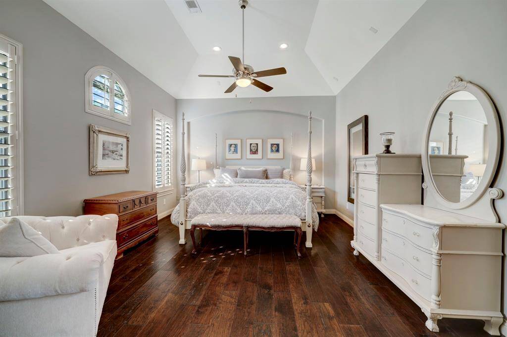 This spacious bedroom features vaulted ceilings, elegant hardwood floors, and abundant natural light through stylish plantation shutters. It includes a cozy sitting area, classic four-poster bed, and ample storage with dressers and nightstands, creating a serene and inviting atmosphere.