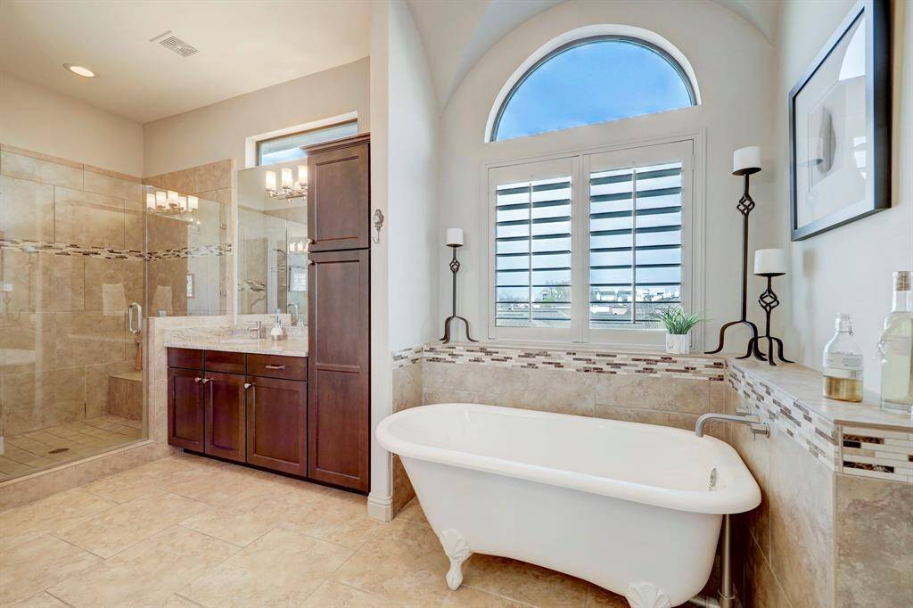 This elegant bathroom features a freestanding clawfoot tub beneath a large arched window, a spacious glass-enclosed shower, and rich wooden cabinetry. It boasts modern fixtures and tasteful tile accents for a luxurious feel.