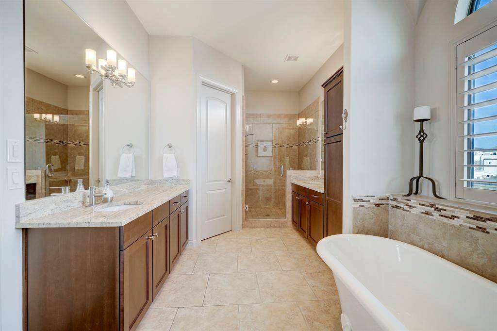 This spacious bathroom features elegant tile flooring, a freestanding bathtub, and a large walk-in shower. The double vanity with granite countertops and dark wood cabinets offers ample storage, complemented by stylish lighting fixtures. Natural light streams in through a window with plantation shutters, creating a bright and inviting atmosphere.