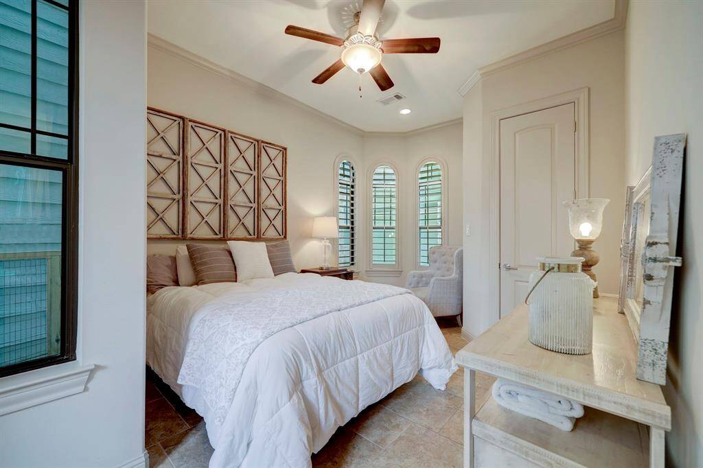 This cozy bedroom features a comfortable bed with decorative pillows, elegant wall art, and ample natural light from three arched windows. A ceiling fan and neutral tones create a relaxing ambiance, complemented by a stylish armchair and a rustic dresser.