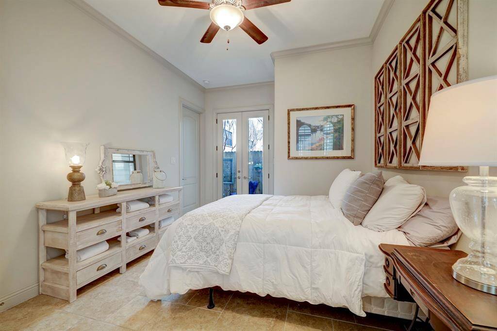 This cozy bedroom features neutral tones, a ceiling fan, and ample natural light through glass doors. It includes a stylish wooden dresser and contemporary decor, creating a warm and inviting atmosphere.