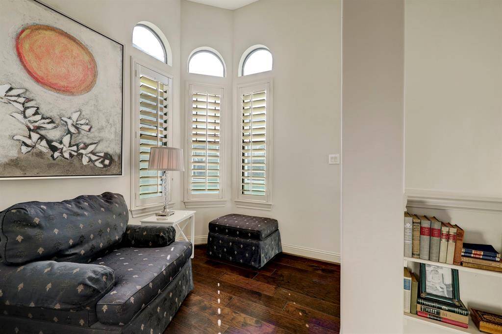 This cozy corner features elegant dark wood flooring, a plush navy armchair, and matching ottoman. The space is brightened by a large window with stylish shutters, complemented by a modern lamp and a colorful abstract painting. Built-in shelving offers additional storage or display space.