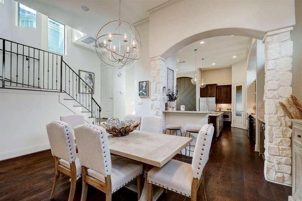 This photo showcases a modern, open-concept dining and kitchen area with elegant lighting, stone accents, and dark wood flooring. A staircase with a metal railing and large windows enhance the space's brightness and sophistication.