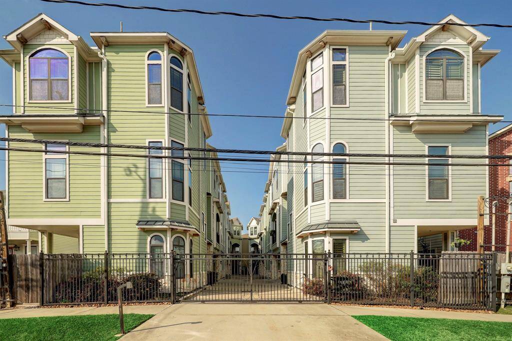 This photo shows a pair of modern, three-story townhouses with light green siding and white trim. They feature multiple windows, a secure gated entrance, and a paved driveway. The townhouses are closely positioned, offering a cozy community feel.