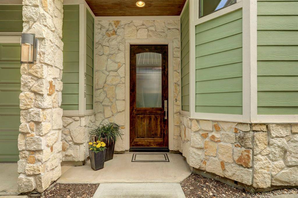 Charming entryway with a rustic wooden door, surrounded by stone and green siding. The covered porch features a modern light fixture and a planter with flowers, offering a welcoming and stylish first impression.