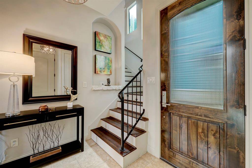 This entryway features a wooden front door with frosted glass, a stylish staircase with dark wood steps and iron railing, and a welcoming console table with a lamp and decor. The walls are adorned with colorful artwork and a large mirror, enhancing the space with light and visual interest.