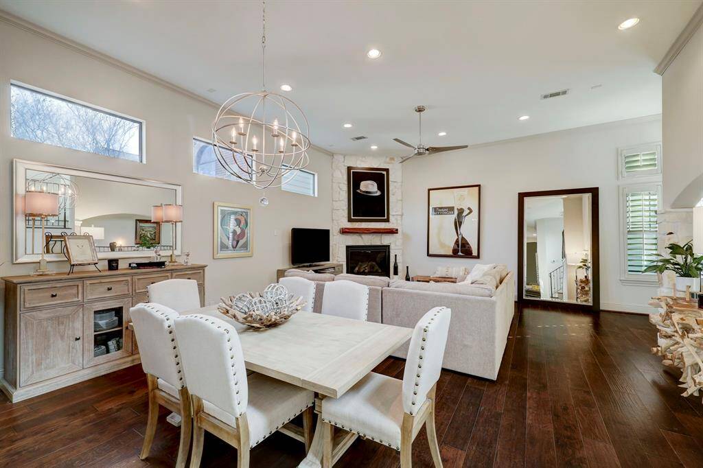 Bright, open-concept living and dining area with high ceilings and hardwood floors. Features a cozy fireplace, modern chandelier, and ample natural light from large windows. Neutral decor creates an inviting and spacious atmosphere.