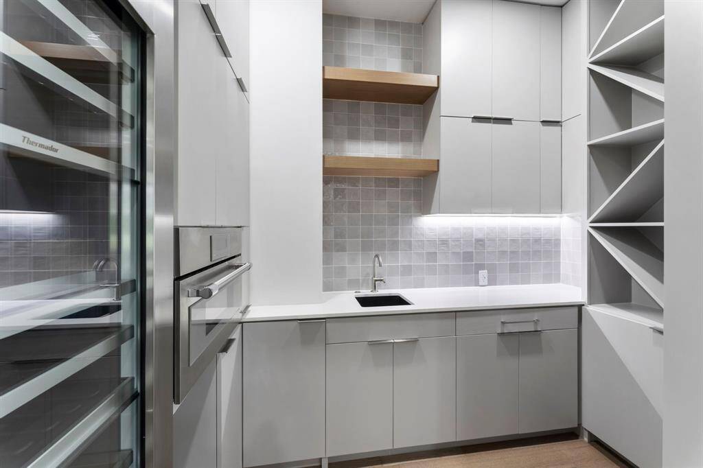 The pantry has a 24" Thermador wine fridge, Thermador microwave, and vegetable sink. It has ample storage, open shelves, and unique shelving units, complemented by under-cabinet lighting and a minimalist design.