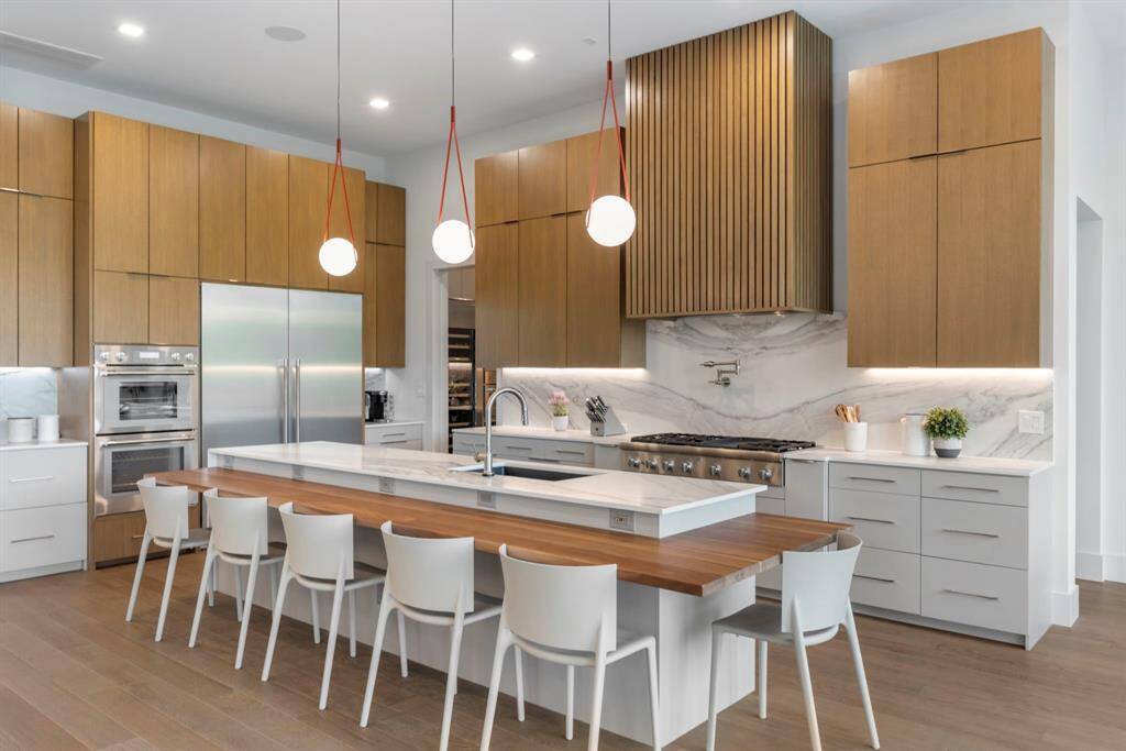 The kitchen features stylish wood cabinetry, a spacious island with seating for six, Thermador stainless steel appliances, and stylish pendant lighting. A quartz matchbook countertop and backsplash and custom designed wooden vent a hood complement the design. The pocket door in the back is the entrance to the pantry. The doorway on the right leads to the primary vestibule.
