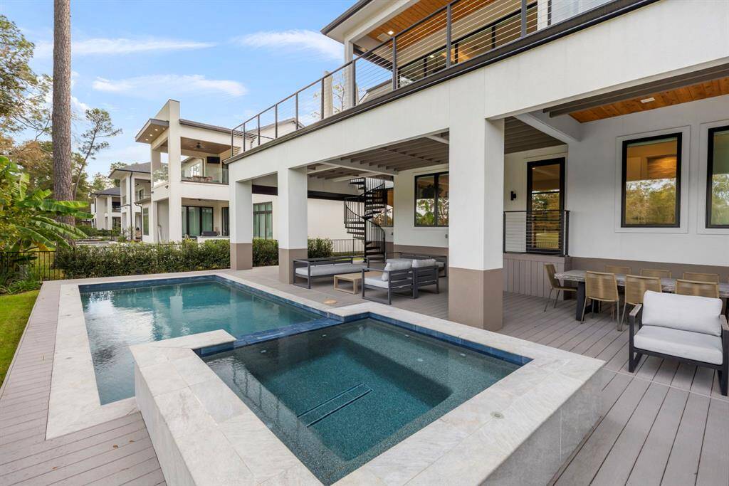 This home features a oversized outdoor deck with a self cleaning pool and hot tub surrounded by lush greenery. The covered patio includes a dining area and spacious seating, perfect for entertaining. A 72" spiral staircase leads to an upper-level balcony, allowing access to both floors.