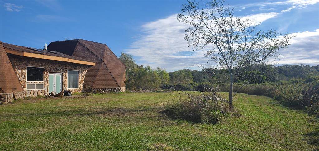 This photo shows a unique geodesic-style home with stone and wood exterior, set in a spacious, grassy yard. It offers a serene, natural setting with open views and a few surrounding trees, perfect for privacy and outdoor enjoyment.