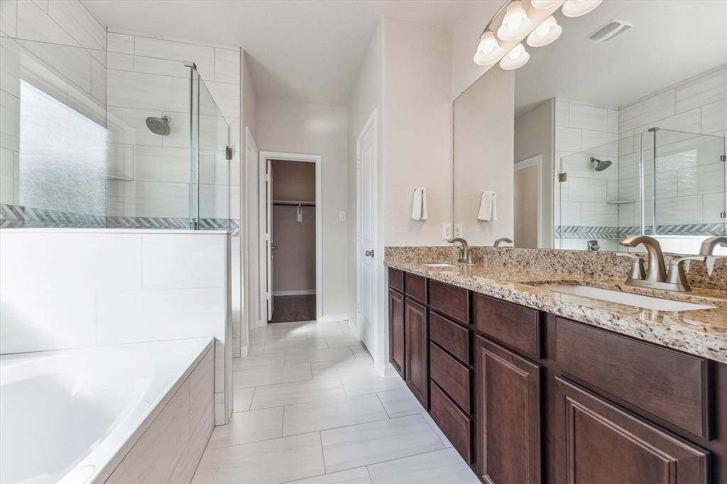 This modern bathroom features a spacious layout with a double vanity and granite countertops. It includes a glass-enclosed shower, a large soaking tub, and a walk-in closet, all accented by sleek tile flooring and contemporary fixtures.