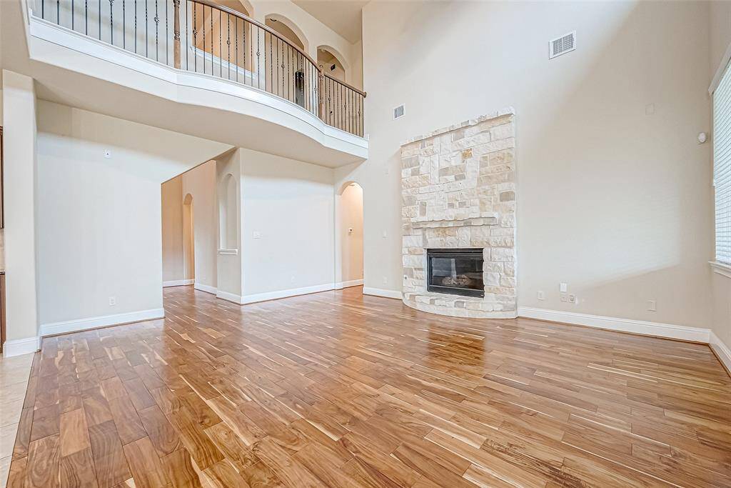 Bright, spacious living room with high ceilings, hardwood floors, and a stone fireplace. It features an elegant staircase and ample natural light from large windows.