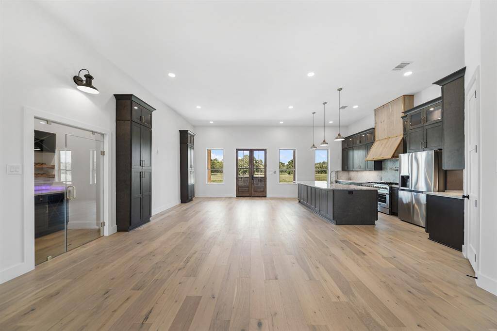 The open-concept kitchen features sleek dark cabinetry, a large island with pendant lighting, and stainless steel appliances. Double doors and windows offer ample natural light, creating a bright and inviting atmosphere.