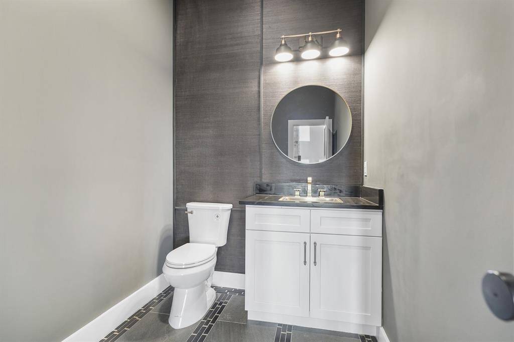 This bathroom features modern design with a sleek black accent wall, round mirror, and contemporary lighting. It includes a white vanity with ample storage and a stylish toilet, complemented by elegant tile flooring.